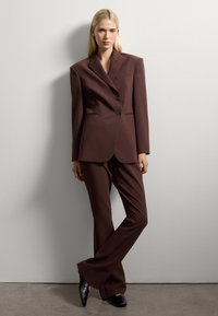 Traje marrón a medida con un blazer estructurado que presenta dos botones, combinado con pantalones acampanados y zapatos de tacón negros. Textura de tela suave.