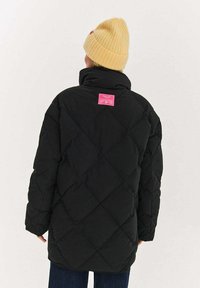 Czarna kurtka puchowa z pikowaniem w kształcie diamentów, wysokim kołnierzem i różową metką z tyłu. Modelka nosi żółtą prążkowaną dzianinową czapkę beanie.