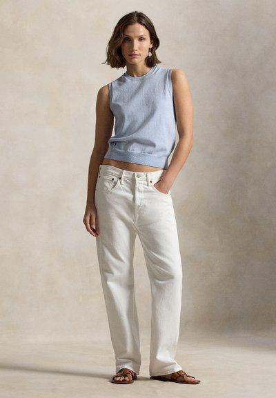 Γυναίκα με ανοιχτόγαλάζιο αμάνικο τοπ, λευκό τζιν ψηλόμεσο και καφέ σανδάλια, που στέκεται μπροστά σε μια μπεζ υφή φόντο.