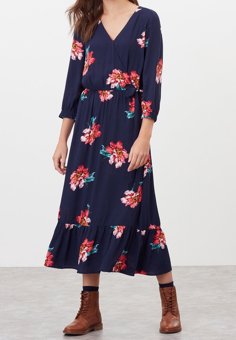 Femme portant une robe midi bleu marine avec imprimé floral rose, manches trois-quarts, ourlet à volants et bottines marron à lacets.