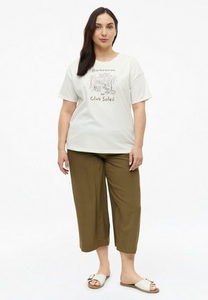 Frau mit dunklen Haaren, die ein weißes T-Shirt mit Grafik und olivgrüne, weit geschnittene, verkürzte Hosen mit weißen Slidesandalen trägt.
