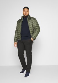 Olive green puffer jacket, navy polo shirt, black pants, and navy sneakers. The jacket features quilted sections and a stand-up collar.
