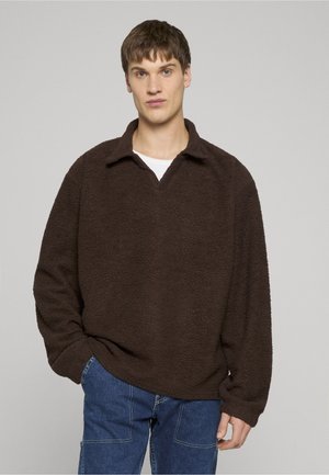 Fiatal férfi rövid barna hajjal, sötétbarna polár pulóvert és kék farmert viselve, sima világosszürke háttér előtt áll.