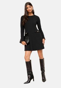 Rochie tricotată neagră cu mâneci lungi, buzunare laterale cu butoane aurii, asortată cu cizme din piele neagră până la genunchi. Design simplu, ajustat.