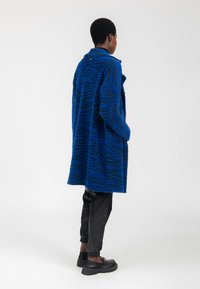 Abrigo texturizado azul con rayas de tigre negras, mangas largas y bolsillos. Combinado con pantalones negros ajustados y zapatos anchos negros.