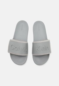 Halli hallid hallid, millel on tekstureeritud rihm, millel on väljaulatuv kiri "Calvin Klein". Sile kummist tald ja pehme jalapadi.