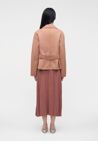 Femme aux longs cheveux noirs portant une veste beige et une jupe midi plissée mauve, debout, de dos, sur un fond uni.