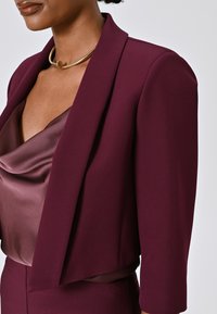 Burgunderfarbener, taillierter Blazer mit drei Viertelärmeln, kombiniert mit einem satinartigen Oberteil mit Cowl-Neck in einem warmen Taupe-Ton, das eine glatte Textur aufweist.