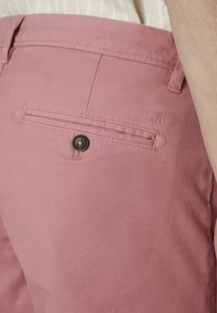 Pink cotton pants featuring a back pocket with a black button and a subtle logo detail stitched onto the fabric. Soft texture and clean lines.