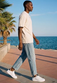 Mies beigen värisessä T-paidassa, vaaleansinisissä farkuissa ja valkoisissa lenkkareissa kävelee merenrantapromenadilla, taustalla palmuja ja valtameri.