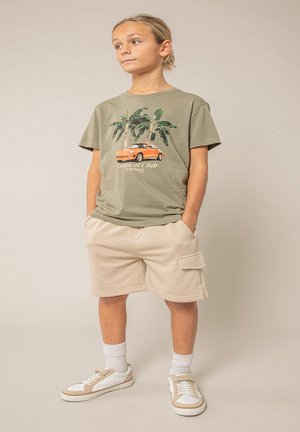 Garçon debout, mains dans les poches, portant un t-shirt vert olive à imprimé tropical, un short beige, des chaussettes blanches et des baskets blanches.