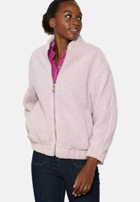 Šviesiai rožinė fliso striukė su užtrauktuku, briaunotu apačia ir šoninėmis kišenėmis. Yra stovinčio apykaklė ir laisvas kirpimas. Lygi, pūkuota tekstūra.