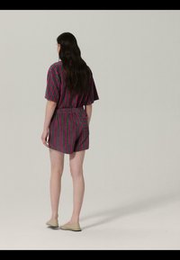 Femme aux longs cheveux noirs portant un ensemble assorti violet et vert à rayures, composé d'un haut à manches courtes et d'un short, se tenant dos à la caméra sur un fond uni.