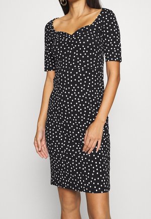 Femme portant une robe noire longueur genou avec des pois blancs et des manches courtes, debout devant un fond clair uni.