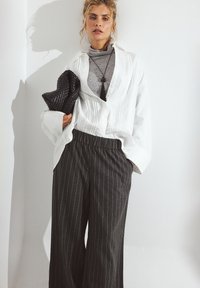 Bela srajca s gumbom in teksturiranim sprednjim delom, siva polo majica, črn pleten clutch in sivo hlače širokega kroja z pinstripi.