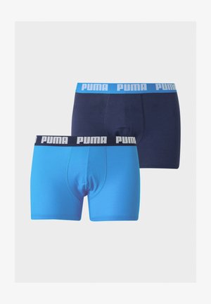 Hele tumenaha nahaga saleikas seisab paljajalu, seljas tumesinised Puma bokserid sinise vöökohaga, vaadates vasakule tavalise tausta ees.
