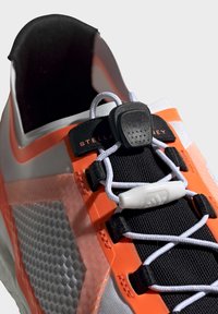 Scarpa sportiva con una tomaia argento, dettagli arancioni e pannello in mesh testurizzato. Caratterizzata da lacci neri e un design elegante e aerodinamico.