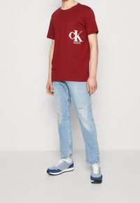 T-shirt rossa in cotone con un logo bianco, abbinata a jeans chiari strappati e sneakers blu con dettagli bianchi e suole nere.