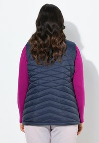 Gilet trapuntato blu navy con motivo a zigzag, indossato sopra un top a maniche lunghe viola. Texture morbida con una finitura leggermente riflettente.