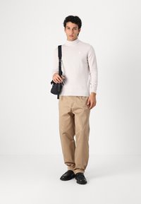 Hellrosa Rollkragenpullover, beige Hose, schwarze Schuhe. Das Model trägt eine kleine schwarze Tasche mit Schulterriemen. Glatte Texturen überall.