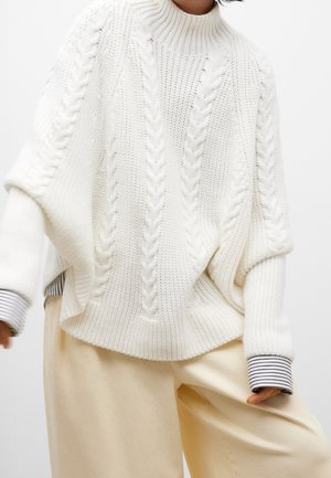 Pull en tricot blanc avec un col montant, présentant des motifs en cable et des manches larges. Associé à un pantalon beige avec des poignets rayés.