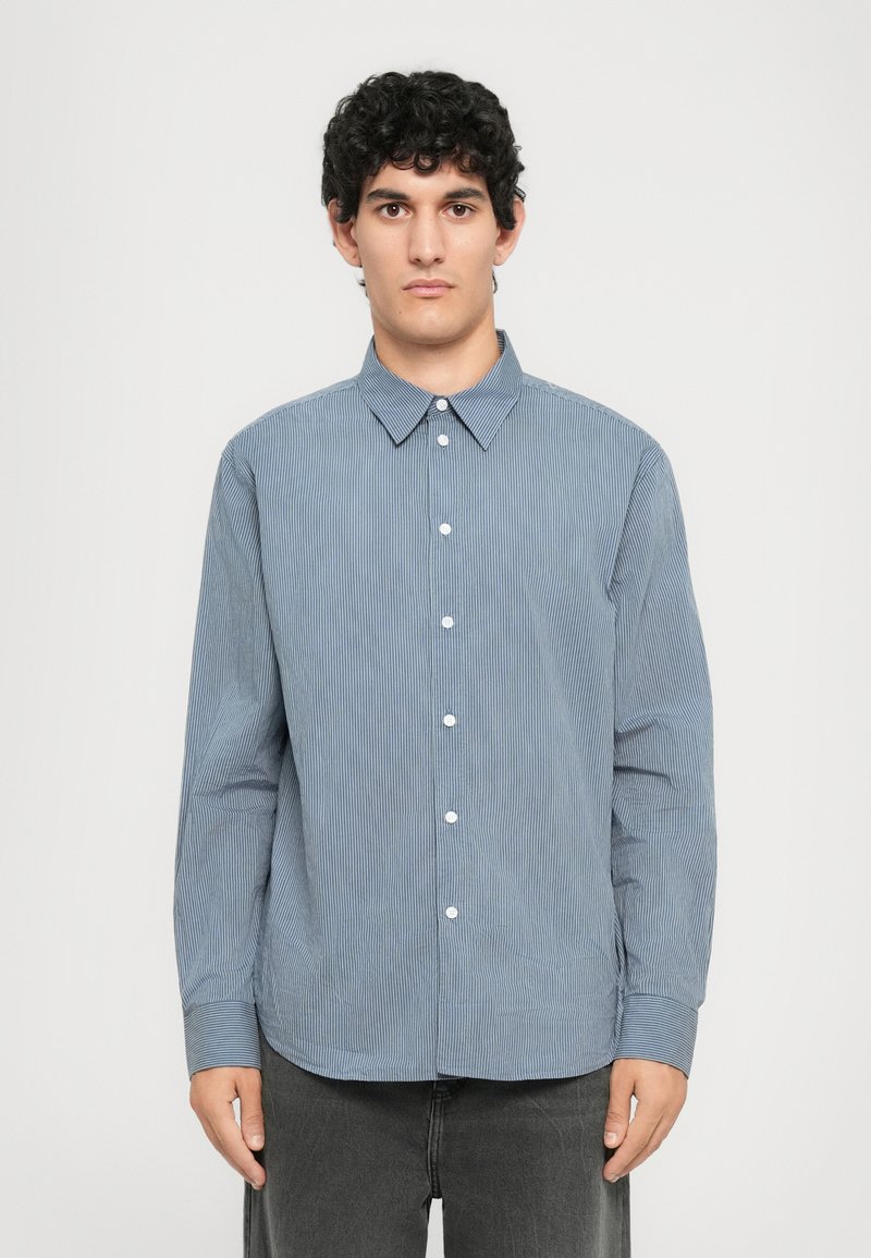 Jeune homme aux cheveux bouclés courts portant une chemise boutonnée rayée bleu et blanc et un pantalon gris foncé, se tenant contre un fond uni.