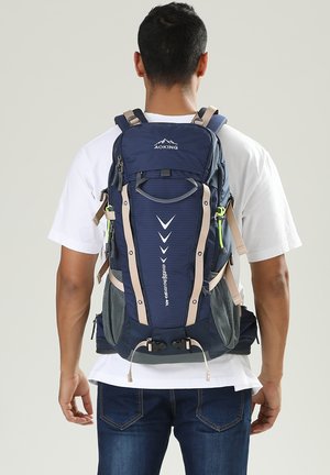 Sac à dos bleu marine avec des sangles beiges, présentant un design texturé, plusieurs compartiments, des poches latérales en mesh et des boucles réglables.