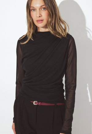 Õlani pikkusega helepruunide juustega naine kannab musta läbipaistvast kangast pika varrukaga särk ja mustad püksid, millel on peen tumepunane vöö.