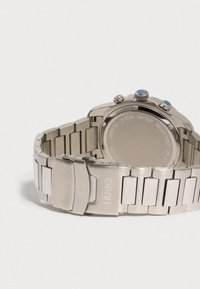 Hopeakuvioinen rannekello, jossa on sinisiä yksityiskohtia painikkeissa, ruostumatonta terästä oleva hihna ja lukko, johon on kaiverrettu "Liu Jo" -logo.