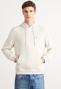 Junger Mann in einem cremefarbenen Hoodie mit einem kleinen "cK"-Logo auf der Brust, die Hände in der vorderen Tasche, kombiniert mit blauen Jeans.