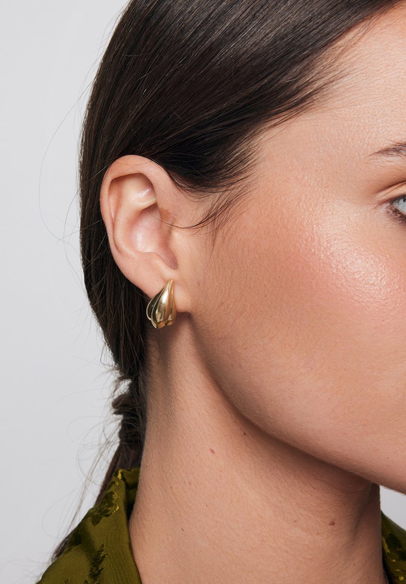 Złote kolczyki w kształcie kropli, przymocowane do płatka ucha, o gładkiej, błyszczącej fakturze i rzeźbiarskim designie, noszone z zieloną bluzką.