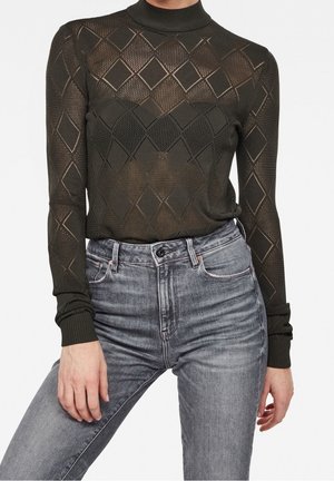 Frau trägt ein schwarzes, transparentes Langarmtop mit Rautenmuster und hoch taillierte ausgewaschene graue Jeans vor schlichtem Hintergrund.
