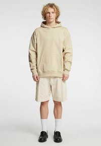 Beige hettegenser med lomme foran, kombinert med lyse beige shorts, hvite sokker og svarte slip-on sko.