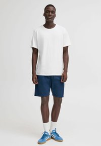 Hvid bomulds t-shirt med rund hals, parret med marineblå shorts. Tilbehøret inkluderer blå atletiske sko og hvide ribbede sokker.