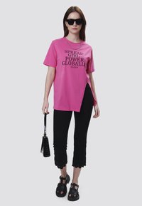 Camiseta rosa de manga corta con texto impreso en negro, combinada con pantalones cortos negros con dobladillos en forma de concha y sandalias negras de suela gruesa.