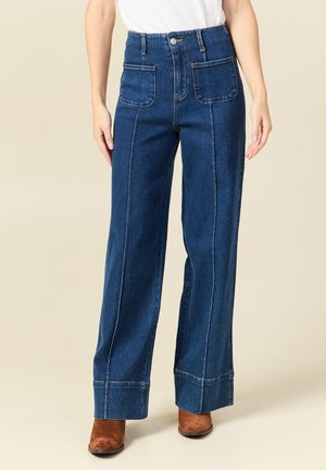 Mujer con vaqueros azules de tiro alto y pata ancha, con bolsillos de parche delanteros, camiseta blanca y botines marrones de ante.