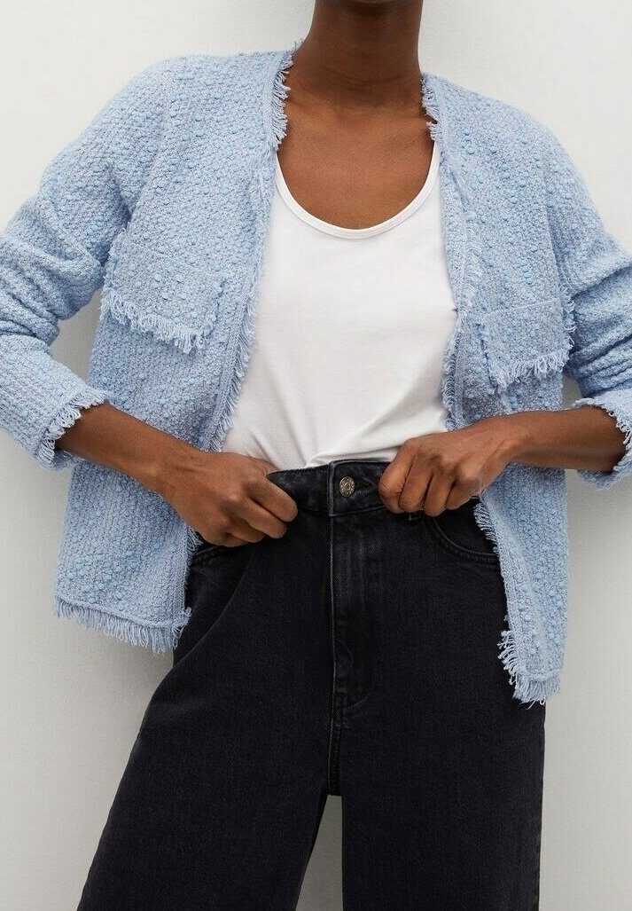 Personne qui ferme un bouton sur un jean noir taille haute, portant un débardeur blanc et un cardigan bleu clair texturé avec des bords à franges.