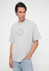 Siva bombažna majica z okroglim izrezom in kratkimi rokavi, ki na prsih nosi krožni logotip z napisom v temneje sivi barvi, centriran.