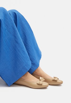 Piernas con pantalones azul brillante y bailarinas beige con lazos decorativos, posadas sobre un fondo blanco liso.