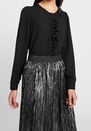 Vrouw draagt een zwarte blouse met lange mouwen en een ruchesdetail aan de voorkant, gecombineerd met een hooggetailleerde metallic zilveren geplooide rok.