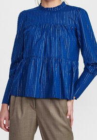 Maglia a maniche lunghe blu con colletto a volant, caratterizzato da strisce verticali, vita arricciata e dettagli testurizzati. Indossata sopra pantaloni marrone chiaro.