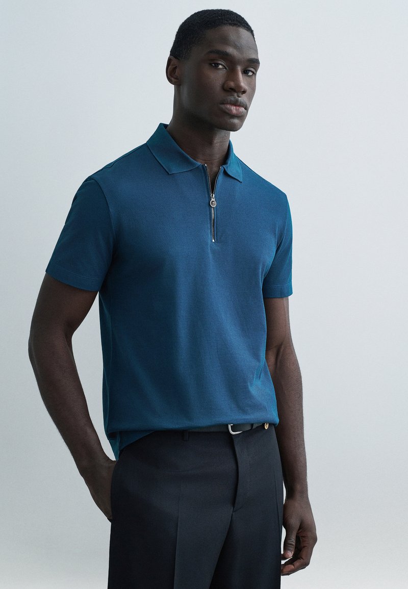 Turkio spalvos polo marškinėliai su trumpomis rankovėmis ir pusiau užtraukiamu apykakle, derinami su tamsiomis kelnėmis. Lygus audinys ir prigludęs dizainas.