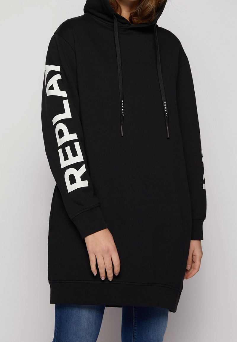 Person som bär en svart lång huvtröja med "REPLAY" i stora vita bokstäver på ärmen och svarta dragsnören med vit text.