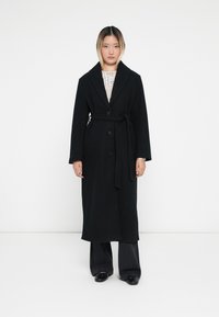 Abrigo de lana negro con solapa ancha, botones y cinta en la cintura, con mangas largas y corte recto, combinado con pantalones negros.