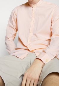 Light peach long-sleeve shirt with a band collar, featuring button closures and a textured fabric. Worn with khaki shorts.