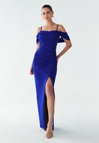 Dolga kraljevsko modra obleka z oprijetim korzetom, odkrito ramo in naborki, z razporkom do stegna. Kombinirana z nevtralnimi sandali z naramnicami.