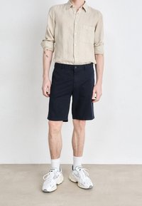 Camicia di lino color beige chiaro con bottoni, pantaloni corti navy in cotone e calzini bianchi con accenti grigi, abbinati a sneakers chunky.