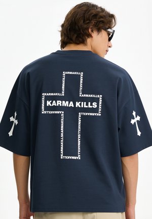 Man draagt een navyblauw oversized t-shirt met een wit kruis en de tekst "KARMA KILLS" op de rug, en witte kruisen op beide mouwen.