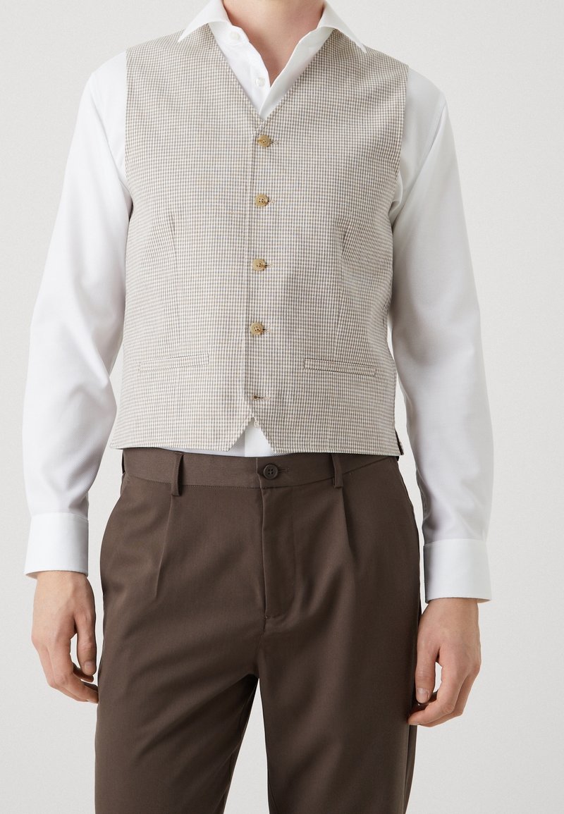 Beige checked waistcoat with five buttons, paired with a white shirt and brown trousers. The fabric is textured with a structured fit.