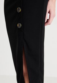 Svart figursydd kjol med en sidoklyv, som har två runda, kontrasterande knappar framtill. Tyget ser slätt och strukturerat ut.